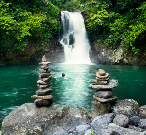 Wasserfall, der in einen klaren Naturpool stürzt, flankiert von gestapelten Steinen im Vordergrund.