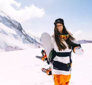 Frau mit Skiklamotten und einem Snowboard in der Hand, im Hintergrund eine schneebedeckte Piste