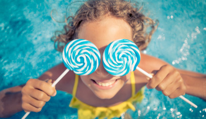 Mädchen mit gelocktem Haar hält zwei blaue Lollis vor die Augen und steht in einem Swimmingpool mit gelbem Badeanzug.